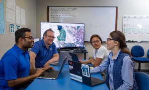 Two men and two women sit in front of computers with a large map in the background.