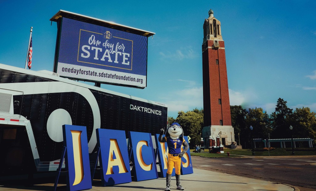 Jack the Jackrabbit poses in front of a sign for One Day for STATE.