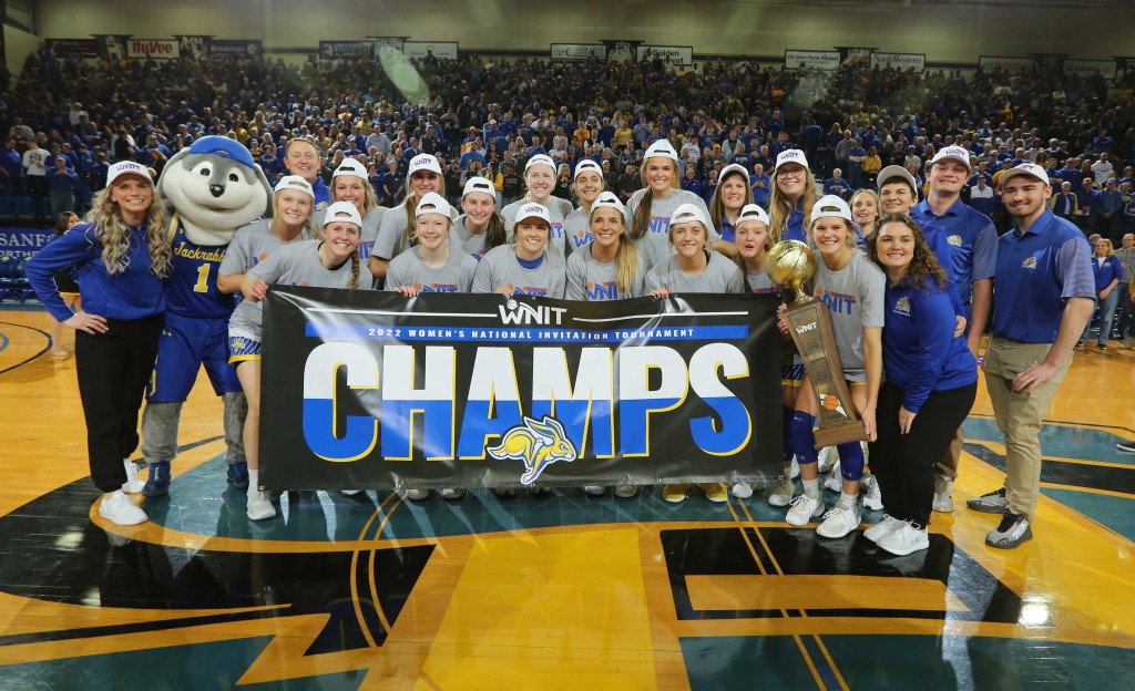 A women's basketball team poses with a tournament trophy and a large banner that says "Champs."