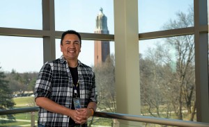 A male college student leans agains a glass window with the Coughlin Campanile in the background.