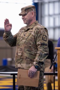 A man in an army uniform stands with his right hand raised.