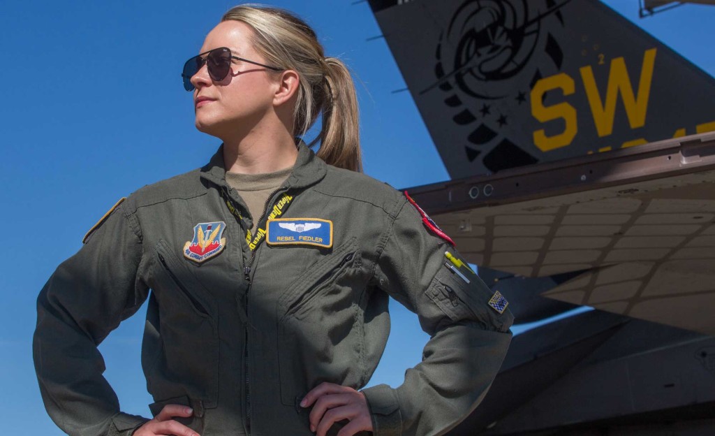 A female fighter pilot stands in front of a jet with her hands on her hips.