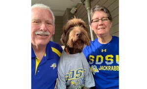 A man and women pose with a brown dog wearing a gray jackrabbits T-shirt.