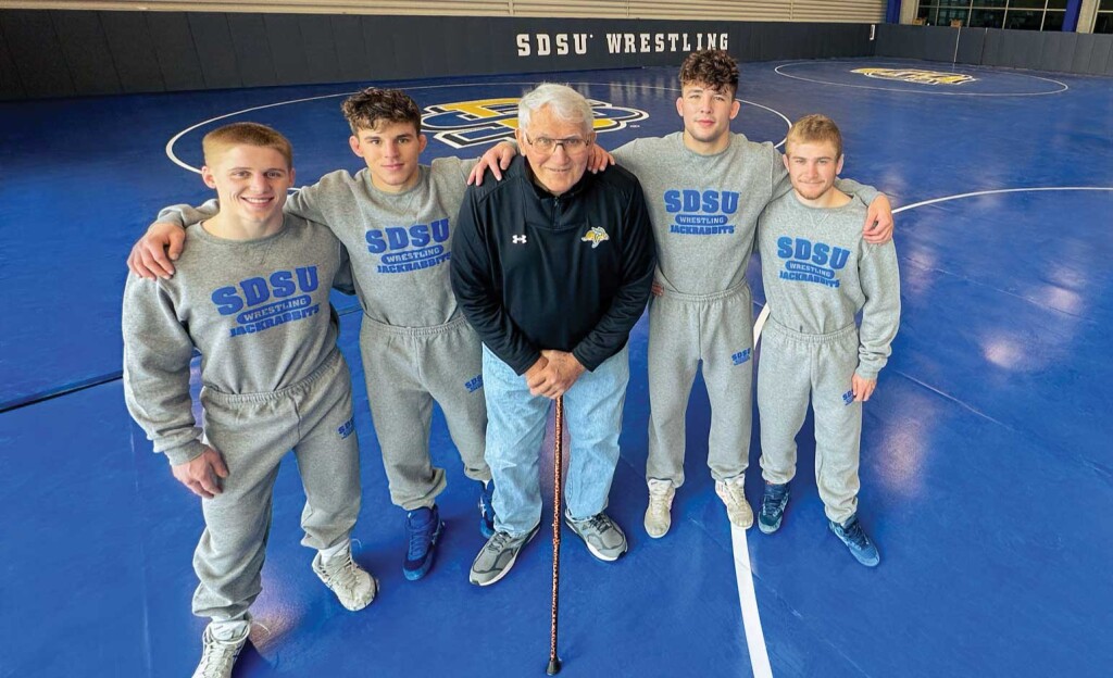 A older man with a cane stands in the middle of four young wrestlers on blue mats.