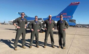 Four military pilots wearing olive green jump suits stand with hands on their hips in front of a military jet. 