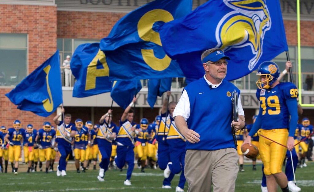 Football coach walks on the field with cheerleaders running behind him waving giant flags.