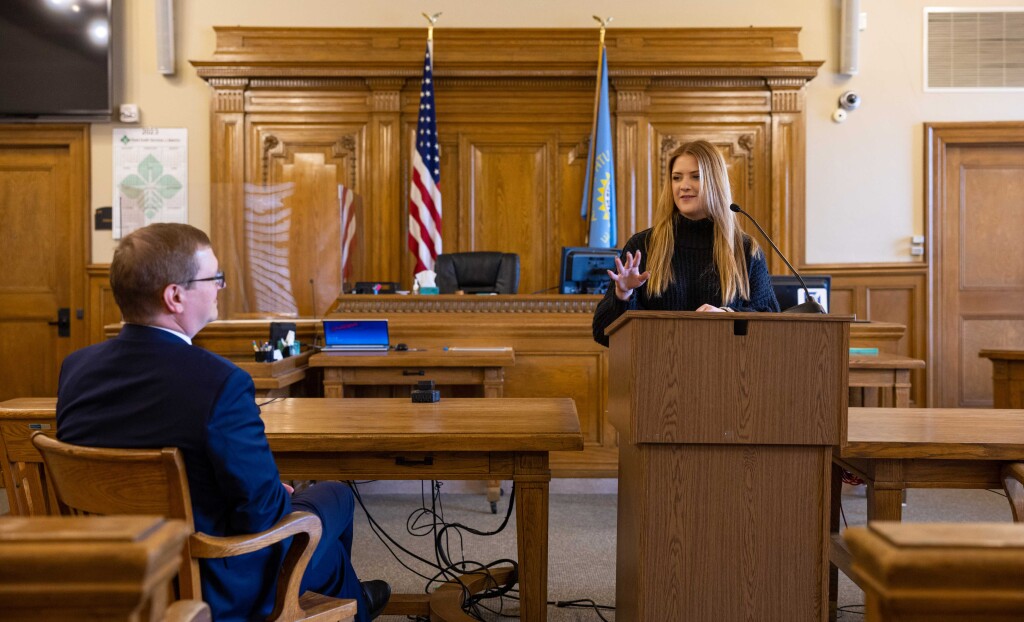 A female college student stands at a podium in a courtroom while a lawyer looks on giving pointers.