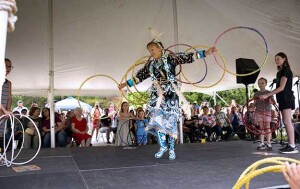 A Native American hoop dancer wears traditional attire and a small head dress and dances with several hoops around their body.