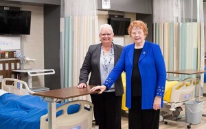 Two women stand in a class room that looks like a hospital room.