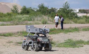 The NASA robot works in a gravel pit while two men watch in the distance.