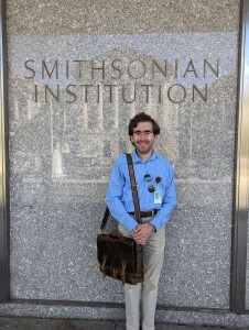 A man with a leather messenger bag on his shoulder stands in front of a stone wall that says Smithsonian Institution.