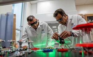 Two men wearing dark glasses and lab coats work with a laser.