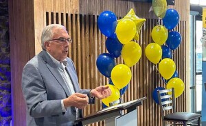 A man speaks at a podium with blue and yellow balloons in the background.
