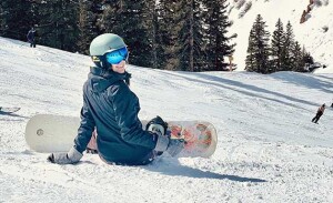 A woman sits on a ski hill with a snowboard strapped to her feet.